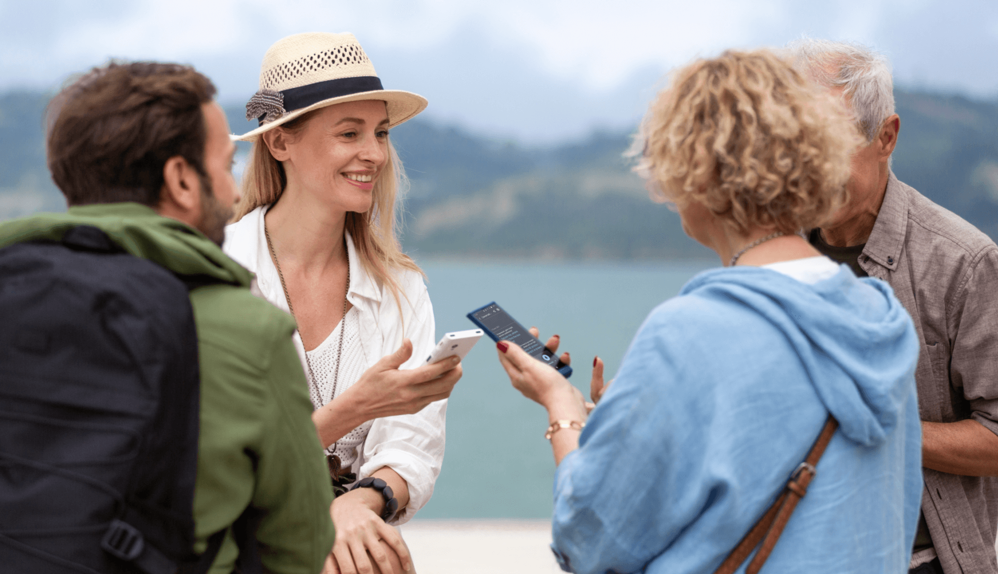 Grupa czterech uśmiechniętych osób rozmawia na tle jeziora lub morza i gór w oddali. Dwie osoby trzymają w dłoniach elektroniczne tłumacze marki Vasco, co sugeruje, że prowadzą rozmowę w różnych językach. Scena ma charakter wakacyjny i towarzyski.
