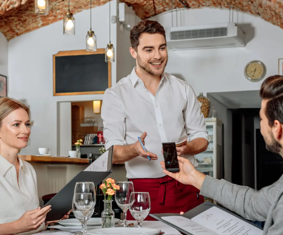 Uśmiechnięty kelner stoi przy stoliku w restauracji, podczas gdy siedzący klient płci męskiej trzyma w ręku tłumacza. Kobieta siedzi obok, trzymając menu. Stół jest zastawiony szklankami, talerzami i małym wazonem z kwiatami.
