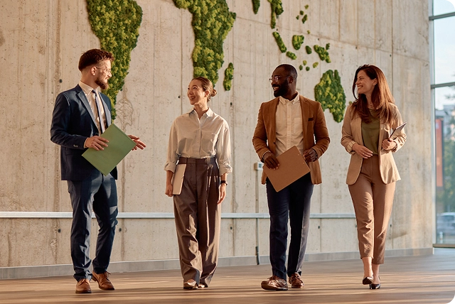 Four smiling people with briefcases walking through a modern office lobby, each wearing a Vasco Translator E1 in their ear. They appear relaxed and confident, engaged in conversation, with a professional and friendly atmosphere.