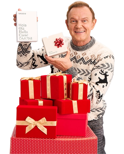 Cezary Pazura stojący za prezentami, w zimowym swetrze, trzyma w rękach pudełko z Vasco Translatorem.