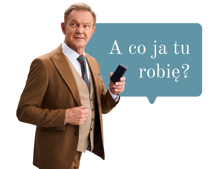 Cezary Pazura w brązowym garniturze, trzymający w ręce Vasco Translator Q1. W tle dymek z tekstem: A co ja tu robię?