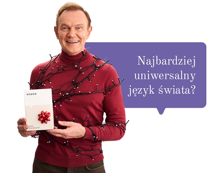 Uśmiechnięty Cezary Pazura w czerwonym swetrze, owinięty lampkami, trzyma w ręce pudełko Vasco Translatora. W tle dymek z tekstem: Najbardziej uniwersalny język świata?