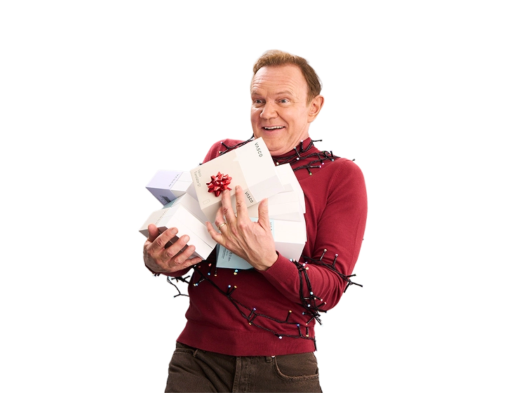 Uśmiechnięty Cezary Pazura ma ręce pełne pudełek z Vasco Translatorami. Ubrany jest w czerwony świąteczny sweter i owinięty lampkami.