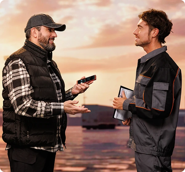 Dwóch mężczyzn rozmawia ze sobą; kierowca ciężarówki trzyma Vasco Translator V4, a osoba stojąca naprzeciwko, prawdopodobnie pracownik magazynu, trzyma dokumenty. Komunikują się za pomocą Vasco Translatora.