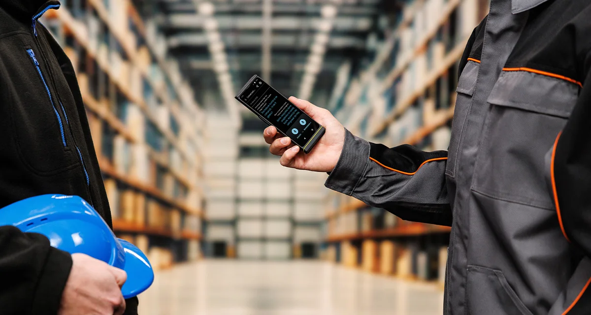 Dwie osoby stoją naprzeciwko siebie; jedna trzyma hełm, a druga Vasco Translator V4. Na ekranie urządzenia widoczny jest aktywny interfejs tłumaczenia mowy.