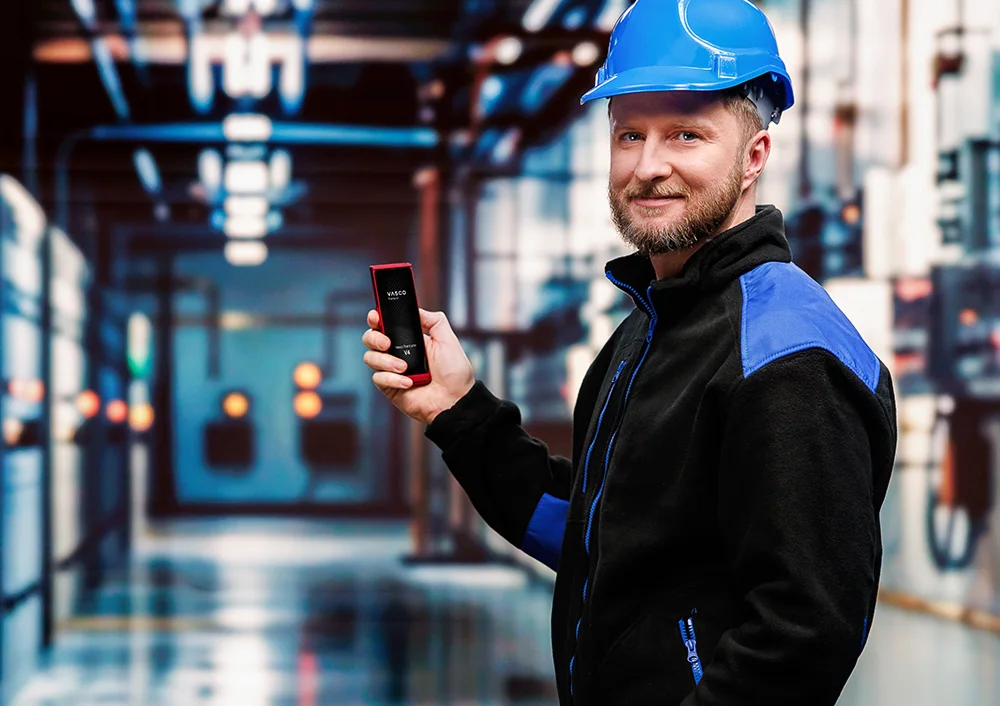 Kierowca ciężarówki stojący przed swoją ciężarówką, trzymający w jednej ręce dokumenty, a w drugiej urządzenie Vasco Translator V4, na którego ekranie widoczna jest funkcja tłumaczenia zdjęć.