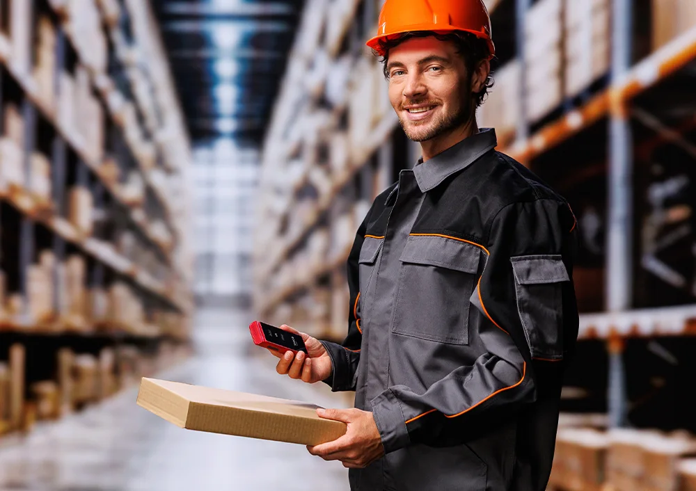 Pracownik magazynu w stroju roboczym i kasku, trzymający w jednej ręce urządzenie Vasco Translator Q1, a w drugiej paczkę, pokazujący dokumenty/etykiety na paczce.