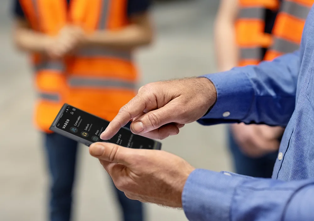 Osoba trzymająca urządzenie Vasco V4, a w tle widać ludzi w strojach roboczych.