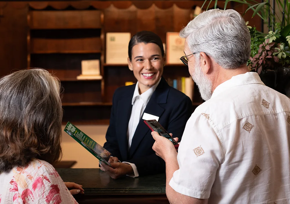 Uśmiechnięta recepcjonistka słucha tłumaczenia z Vasco Translator V4 trzymanego przez mężczyznę zwróconego w jej stronę.