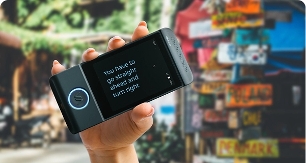 Zbliżenie na dłoń trzymającą czarny tłumacz elektroniczny marki Vasco w pozycji poziomej. Ekran jest w trybie pełnoekranowym i wyświetla przetłumaczony tekst dużą, wyraźną czcionką w kolorze niebieskim: „Musisz iść prosto, a potem skręcić w prawo”. W tle widoczna jest żywa, rozmyta scena plenerowa z kolorowym słupkiem pokrytym znakami kierunkowymi wskazującymi różne kraje, w tym Polskę, Chile i Danię. Jasne światło słoneczne podkreśla tekst na urządzeniu, ułatwiając jego odczytanie. 