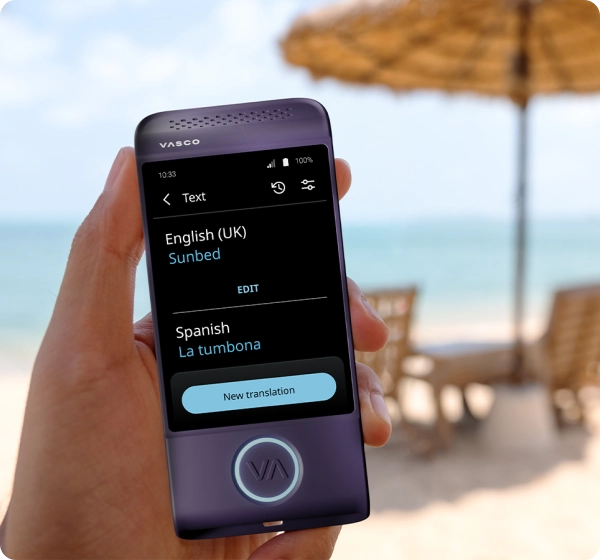 Zbliżenie na dłoń trzymającą fioletowe urządzenie tłumaczące marki Vasco. Na ekranie widnieje tłumaczenie tekstu z języka angielskiego (brytyjskiego) na hiszpański, w którym słowo „Sunbed” zostało przetłumaczone jako „La tumbona”. W tle widać jasną, słoneczną plażę z zamazanym obrazem oceanu, złotym piaskiem, kilkoma leżakami i słomkową parasolką plażową. 