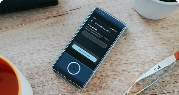 Ujęcie z góry, zbliżenie na ciemnoniebieski tłumacz elektroniczny marki Vasco w przezroczystym etui ochronnym, leżący na jasnym drewnianym stole. Ekran urządzenia jest aktywny i wyświetla menu „Udostępnianie rozmowy” z polem do wpisania adresu e-mail oraz przyciskiem „Udostępnij rozmowę”. Wokół urządzenia na stole widoczne są krawędzie pomarańczowego kubka do kawy, biała doniczka oraz biała smycz z nadrukiem „Hej!”. Scena jest jasno oświetlona, co tworzy czystą i nowoczesną estetykę. 