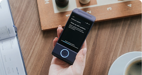 Ujęcie z góry przedstawiające dłoń osoby trzymającej fioletowy tłumacz elektroniczny marki Vasco nad drewnianym stołem. Na ekranie urządzenia widnieje komunikat „Aktualizacja oprogramowania” z napisem „Bądź na bieżąco z aktualizacjami” oraz pasek postępu z tekstem „Pobieranie aktualizacji...”. Na stole wokół urządzenia leżą drewniana szachownica z figurami, otwarty kalendarz oraz biała filiżanka kawy. Oświetlenie jest miękkie i naturalne, tworząc swobodną, codzienną atmosferę. 