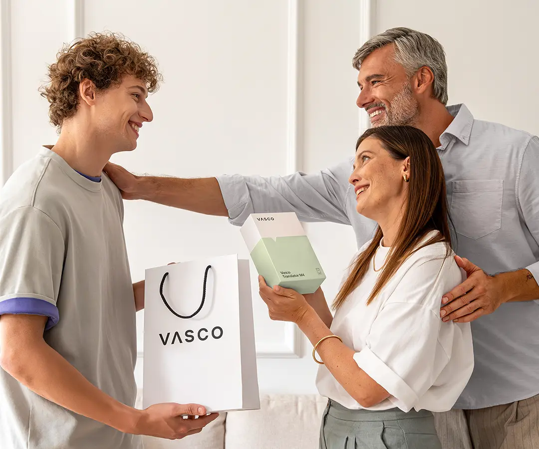 Wzruszająca scena przedstawiająca trójkę uśmiechniętych osób stojących razem. Po lewej stronie młody mężczyzna o kręconych włosach trzyma białą torbę na zakupy z napisem „VASCO”. Pośrodku kobieta trzyma biało-jasnozielone pudełko z napisem „Vasco Translator M4” i patrzy radośnie na młodego mężczyznę. Za nią stoi mężczyzna z siwymi włosami, uśmiechający się ciepło, z jedną ręką na ramieniu kobiety, a drugą na ramieniu młodego mężczyzny. Znajdują się w jasnym pomieszczeniu z neutralnymi ścianami i sofą w tle. 