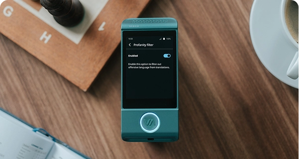 Ujęcie z góry przedstawiające turkusowy tłumacz elektroniczny marki Vasco, leżący na drewnianym stole. Ekran urządzenia jest włączony i wyświetla menu ustawień „Filtr wulgaryzmów”, w którym przełącznik jest ustawiony w pozycji „Włączone”. Pod przełącznikiem widnieje opis: „Włącz tę opcję, aby odfiltrować obraźliwe słowa z tłumaczeń”. Urządzenie otoczone jest przedmiotami codziennego użytku, w tym drewnianą szachownicą z ciemnymi figurami, rogiem otwartego notatnika i białą filiżanką do kawy, a wszystko to uchwycone w miękkim, naturalnym świetle. 
