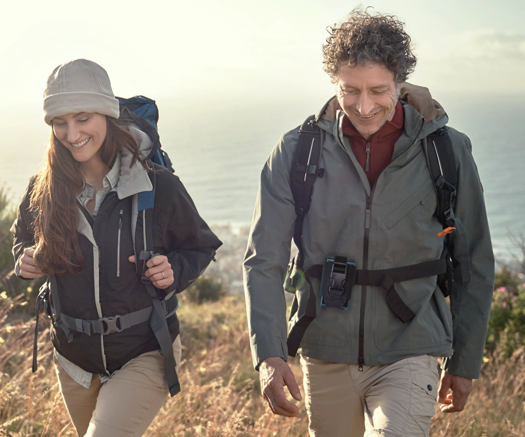 Mężczyzna i kobieta wędrują pod górę trawiastą ścieżką z oceanem w tle. Oboje mają na sobie kurtki i plecaki turystyczne, wyglądają na szczęśliwych i zrelaksowanych w słońcu. Mężczyzna ma przypięty do paska wytrzymały pokrowiec marki IDOGEAR na translator Vasco, w którym znajduje się Vasco Translator Q1.