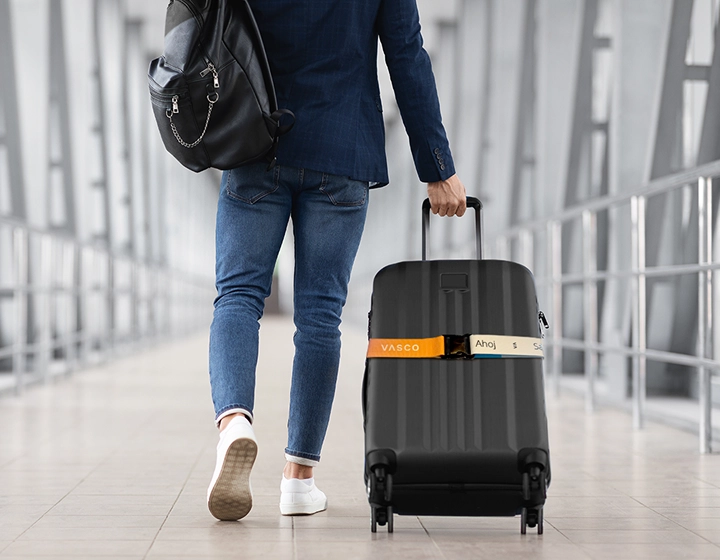 Black suitcase with a rainbow Vasco security strap being pulled by a man walking through an airport.